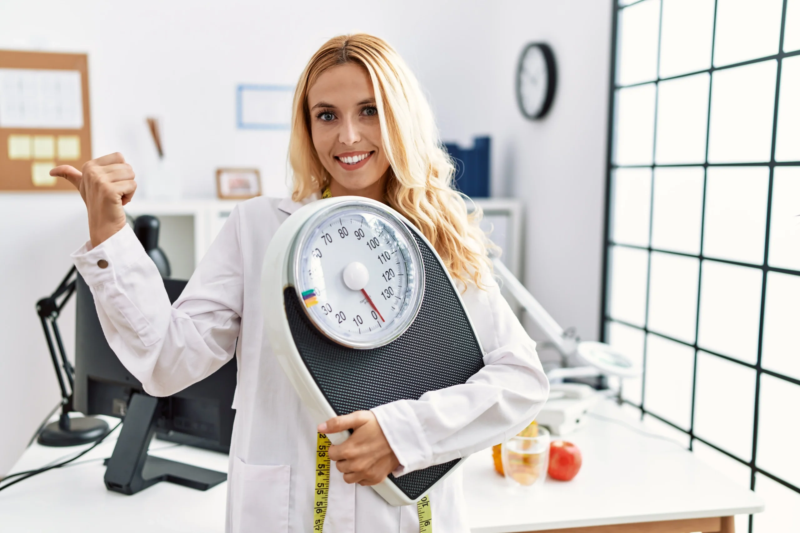 beautiful blonde nutritionist woman holding weight machine balance weight loss pointing thumb up side smiling happy with open mouth (1) 11zon
